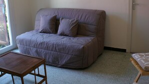 Ein Zimmer mit einer Couch, einem kleinen Tisch und einem Sessel. Eine Tür und ein Fenster sind vorhanden.