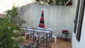 Un patio con sillas de plástico blanco y una mesa, una sombrilla a rayas y una parrilla pequeña.