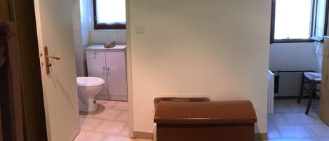 A bedroom with a bed, a wooden chest, a bathroom with a toilet and sink, and a window with a view.