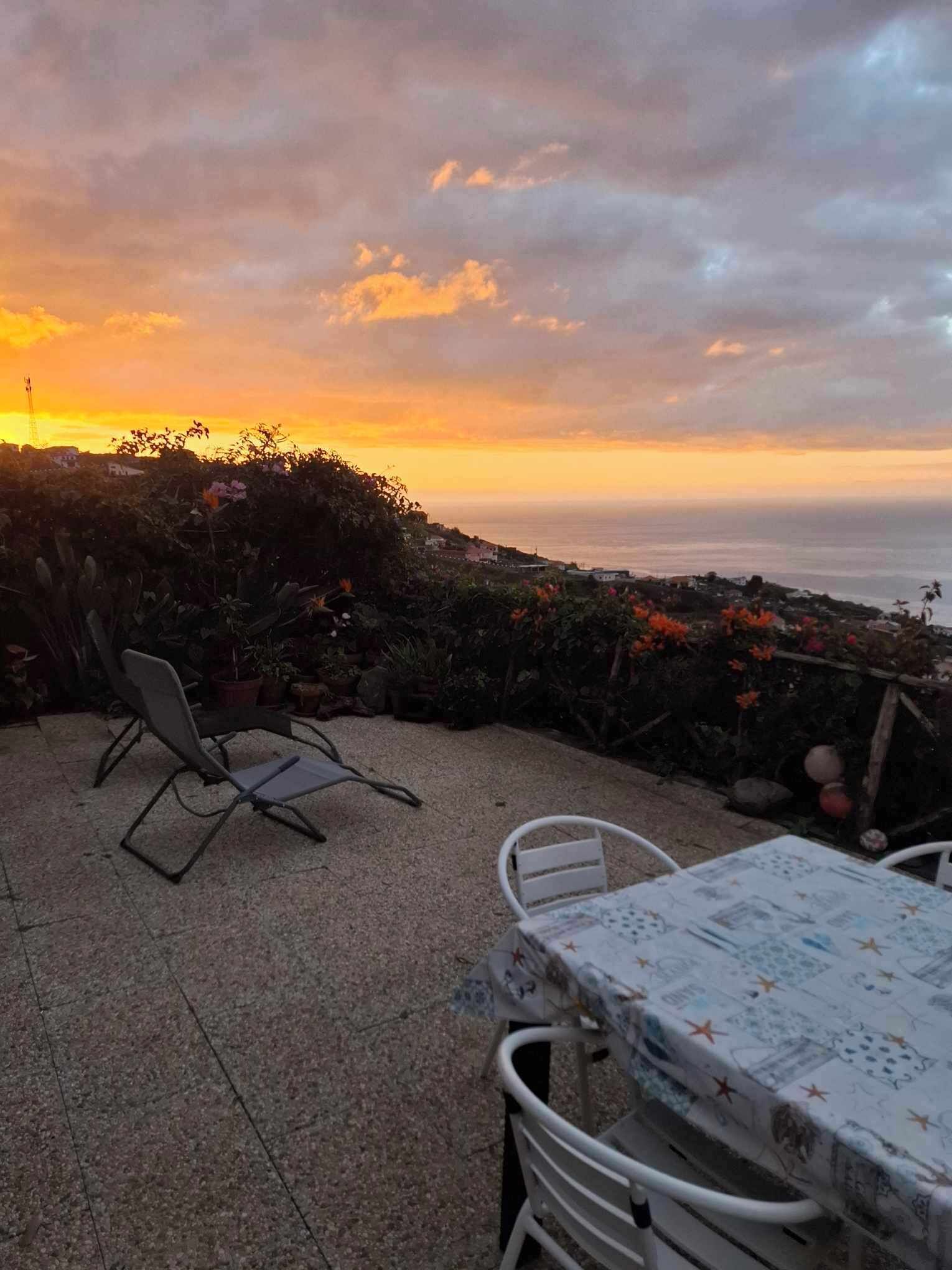 Eine Terrasse im Freien mit Tisch und Stühlen, mit Blick auf eine Küstenlandschaft bei Sonnenuntergang.