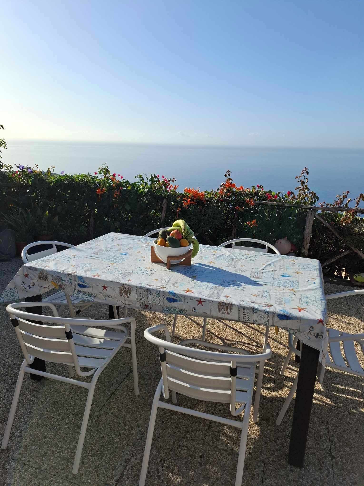 Une table d’extérieur avec un panier de fruits, entourée de chaises blanches, offrant une vue panoramique sur la mer.