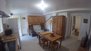 Een ruimtelijk ingerichte leefruimte met kitchenette, een eethoek met een houten tafel en stoelen, een bank en een lamp.