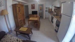 Een compacte leefruimte met een bank, houten kast, eettafel en een keukenhoek met een koelkast en spoelbak.