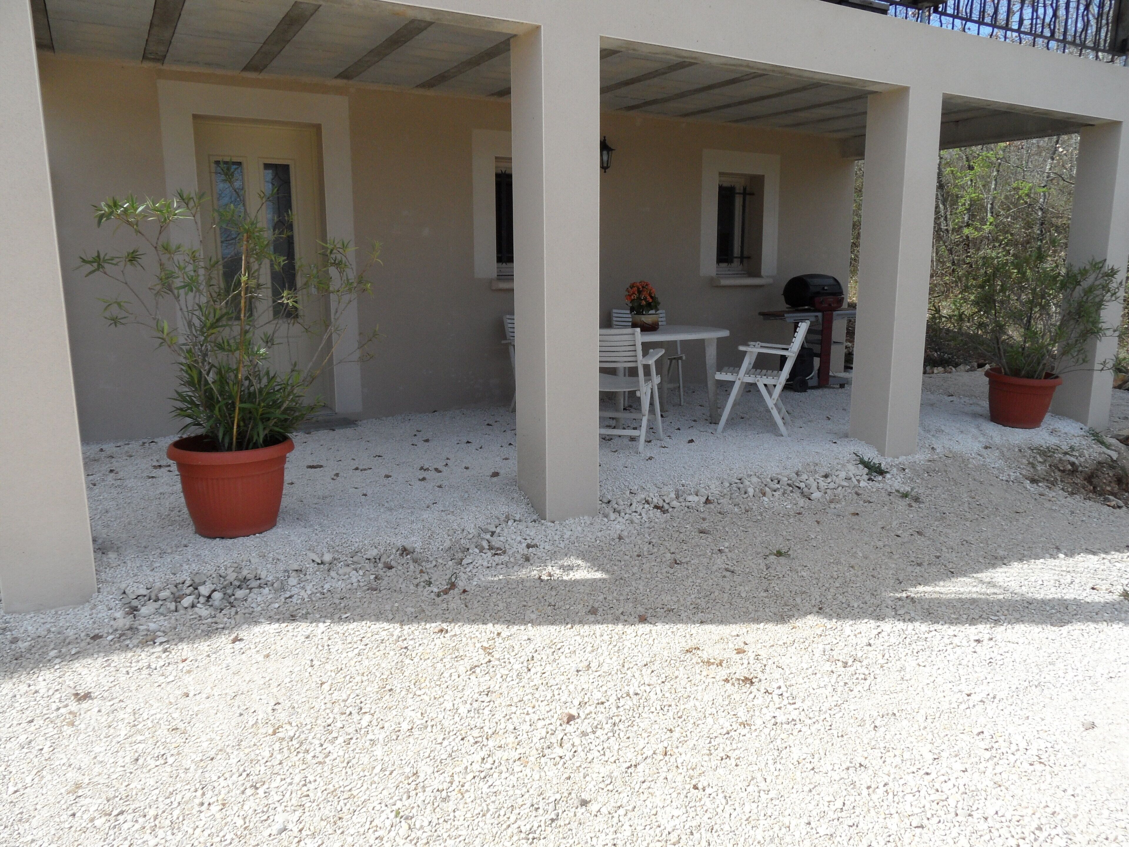 A covered outdoor patio with a table and chairs, surrounded by potted plants and a gravel ground.