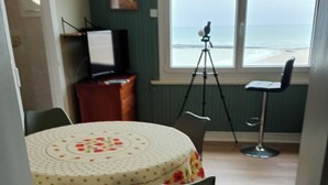 Une pièce avec une table ronde recouverte d’une nappe à motifs floraux, un trépied avec un appareil photo, et une vue sur l’océan par la fenêtre.