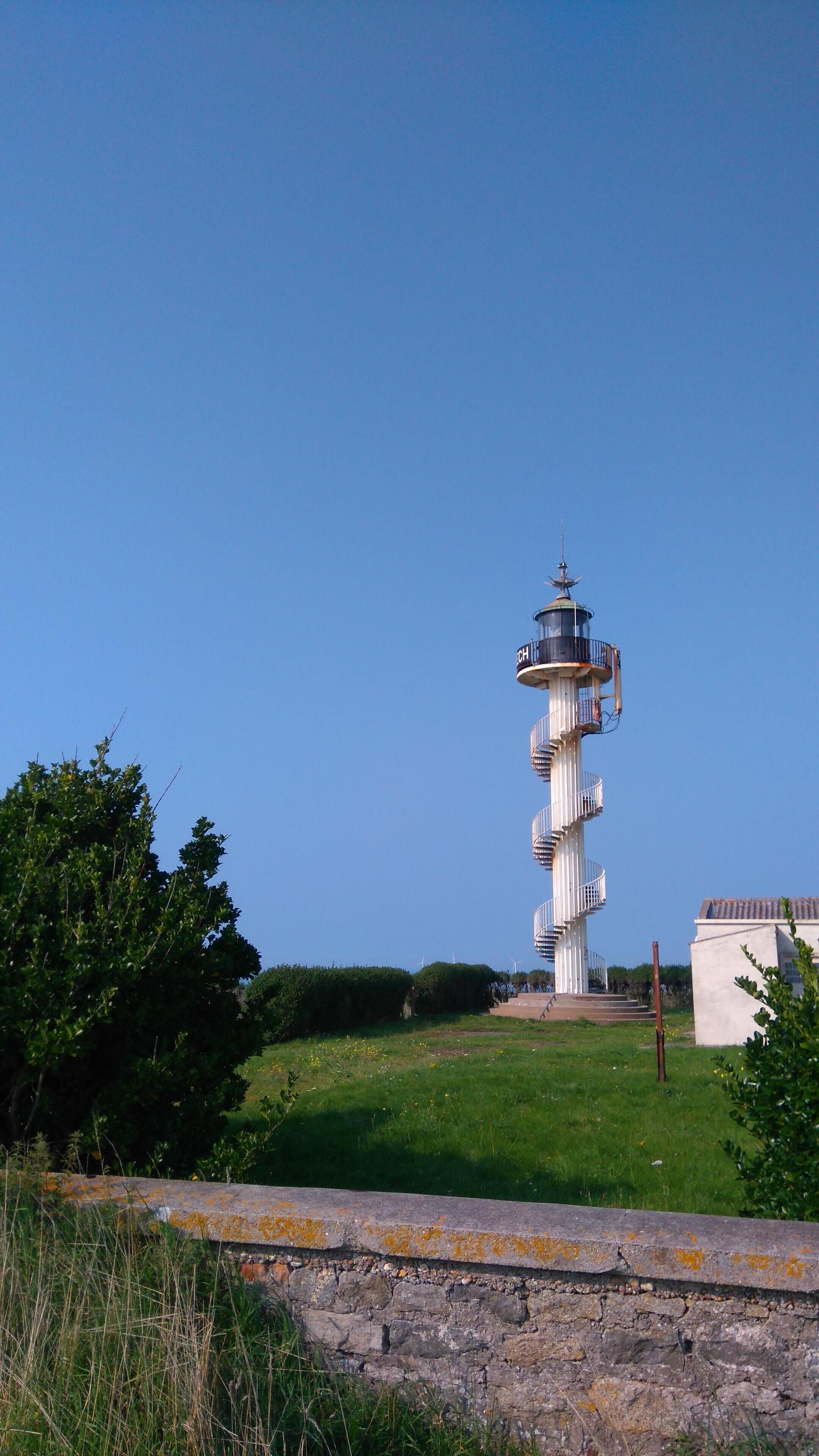 A lighthouse with a spiral staircase, surrounded by greenery and a clear sky.