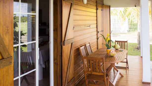 A wooden deck with a table and chairs, a sliding door, and a view of a garden. - Villa Marga: pretty new Creole villa (Capesterre - Belle - Eau)