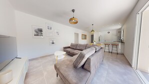 Ein modernes Wohnzimmer mit einer Eckcouch, einem Flachbildfernseher und einem Essbereich, im Hintergrund befindet sich eine Küche.