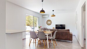 Ein modernes Wohnzimmer mit Essbereich, einem braunen Sofa, einem Fernseher und einem großen Fenster mit Blick auf Grünflächen.