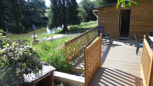 Een houten terras met uitzicht op een rivier en bomen, een houten blokhut met een deur en buitenzitplaatsen.