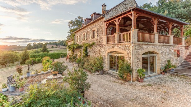 Une maison en pierre dotée d’un balcon, entourée d’une allée gravillonnée et de diverses plantes.