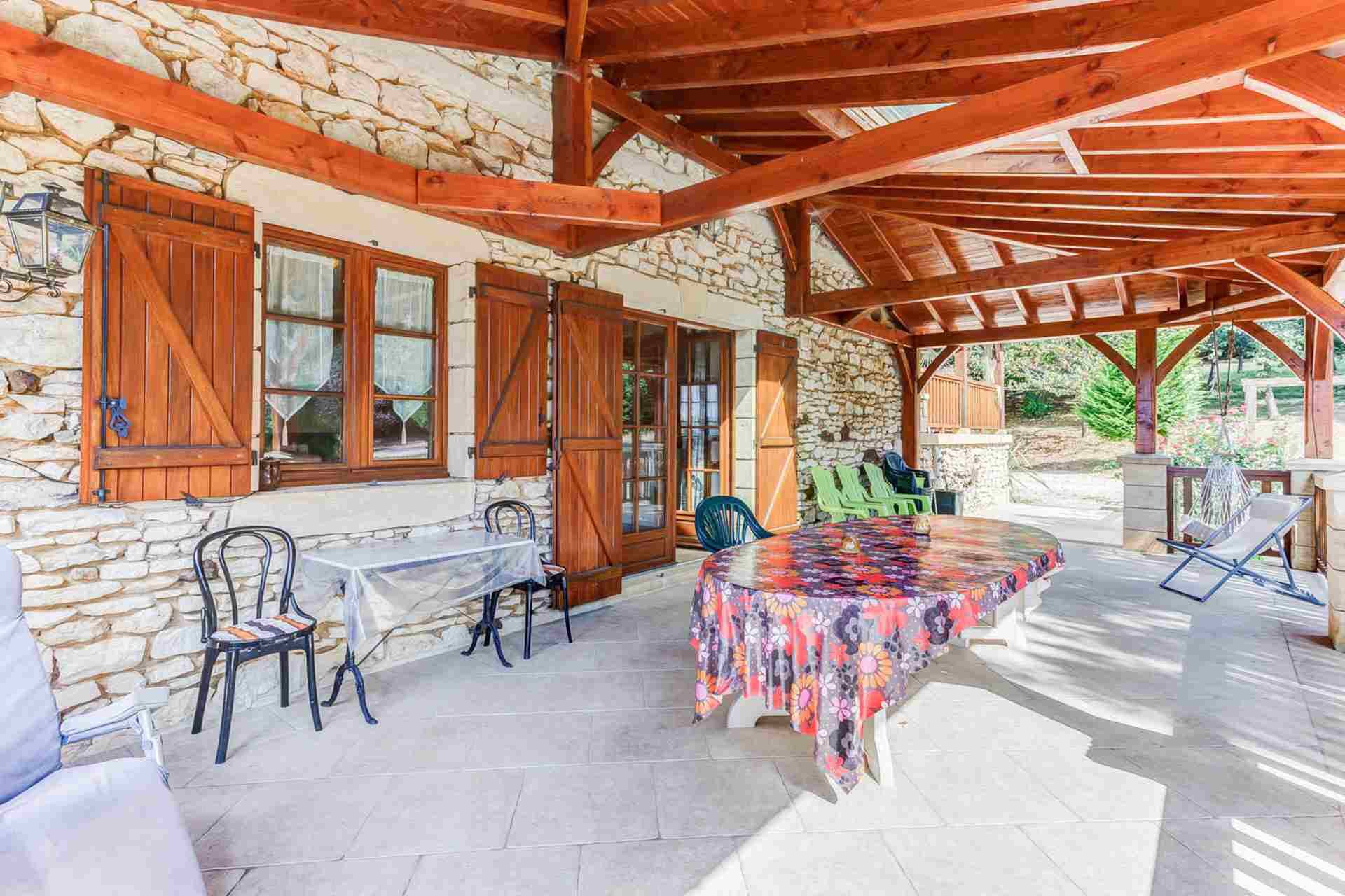 A covered patio with stone walls, wooden beams, a table with a floral tablecloth, and outdoor chairs.