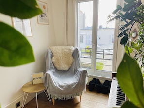 Une pièce chaleureuse avec un fauteuil près d’une fenêtre, une petite table sur laquelle est posé un panneau, et un piano.