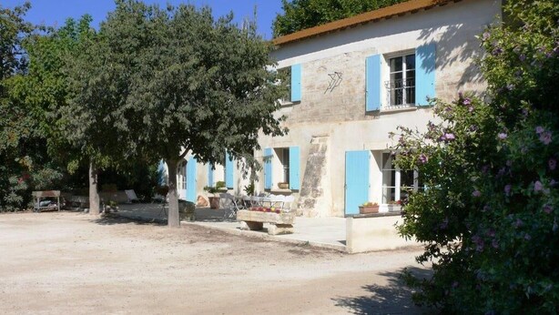 Ein zweistöckiges Gebäude mit blauen Fensterläden, ein Garten mit Bäumen und Blumen sowie eine Bank im Vordergrund.