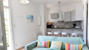 Eine moderne Küche mit weißen Schränken, einer Mikrowelle und einer Kaffeemaschine. Im Wohnbereich befindet sich ein petrolfarbenes Sofa mit bunten Kissen.