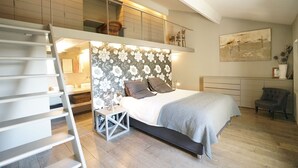 Une chambre à coucher comprenant un lit, une table de chevet, une chaise, un tableau et un espace en mezzanine avec une fenêtre.