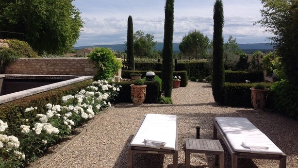 Un jardin avec un chemin de gravier, des rosiers blancs et de grands arbres. On y trouve deux fauteuils de jardin en bois, dotés de coussins blancs.