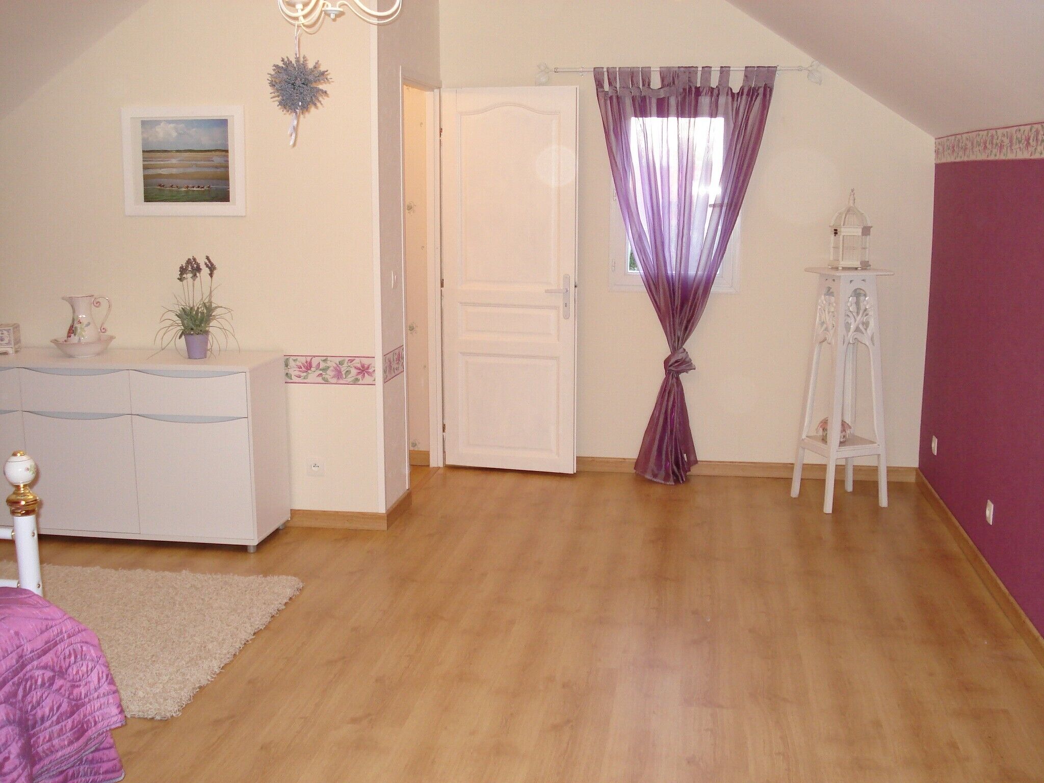 Una habitación con piso de madera, puerta blanca, cortina morada, mueble blanco y una pequeña planta.