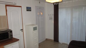 A compact kitchen with a microwave, a small refrigerator, and a cabinet. There is a door with a window, a wall-mounted heater, and a curtain.