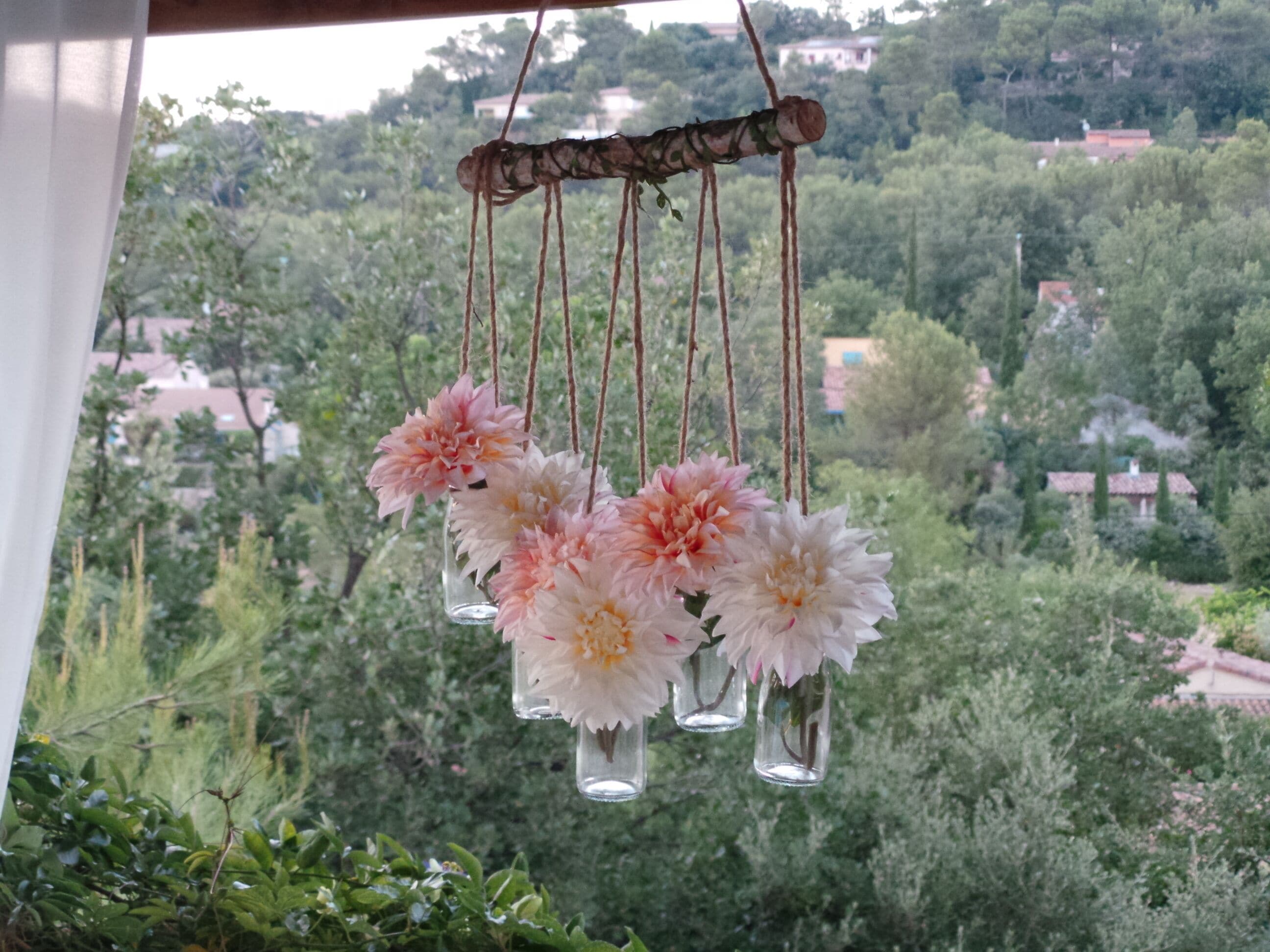 Eine hängende Dekoration mit Blumen in klaren Glasbehältern, vor einer Kulisse aus Grünpflanzen und Gebäuden.