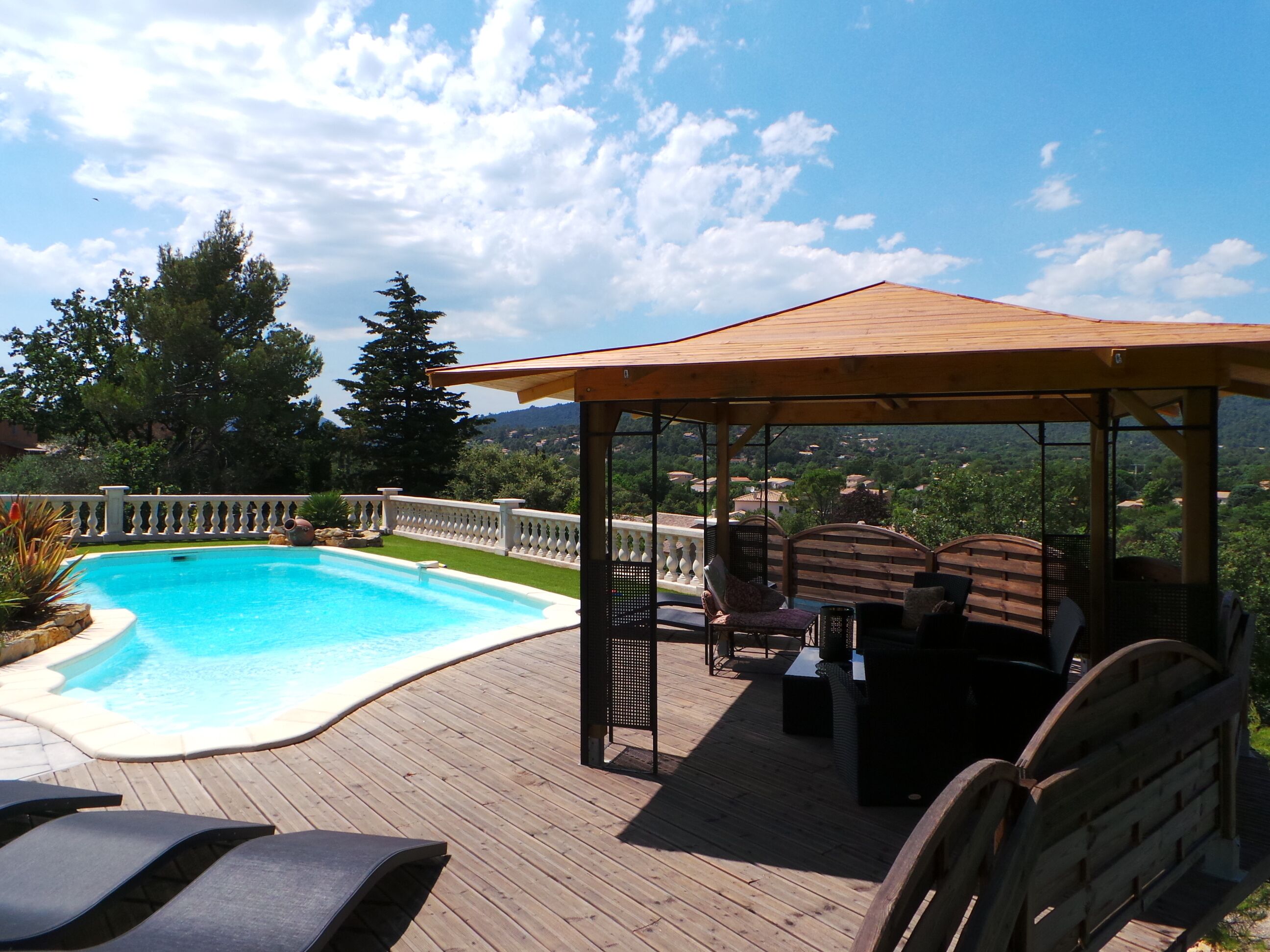En træterrasse med swimmingpool, liggestole og en pergola med udsigt over et naturskønt landskab.