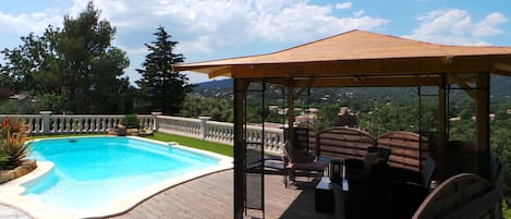 En træterrasse med swimmingpool, liggestole og en pergola med udsigt over et naturskønt landskab.