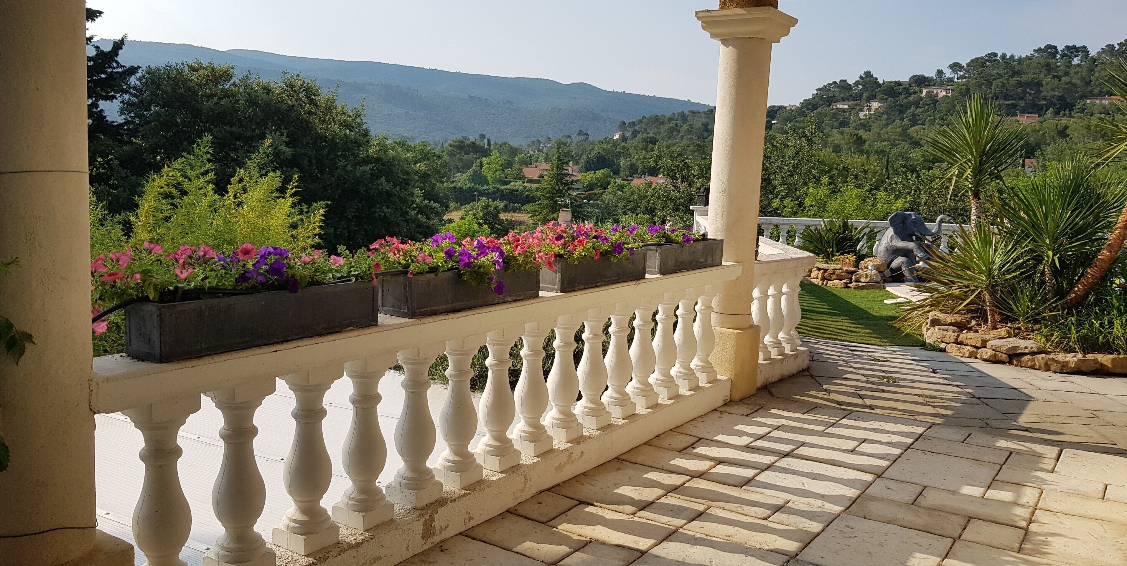 Un balcon doté d’une balustrade blanche, de plantes en pot et offrant une vue sur un paysage verdoyant avec des collines.