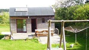 Une petite cabane en bois avec un toit en pente, une terrasse avec une table et des bancs, et un fil à linge dans la cour.