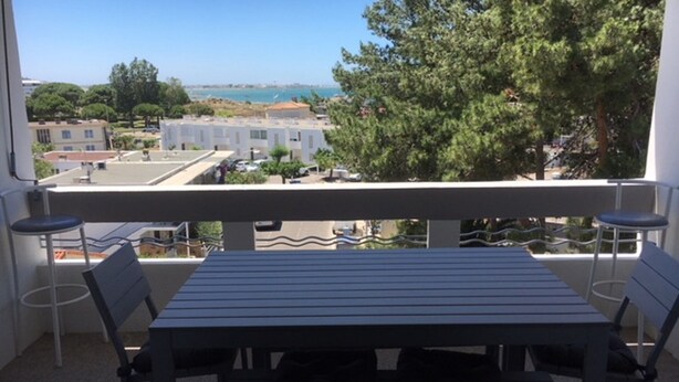 A balcony with a table and chairs, overlooking a residential area with houses and trees. - Apartment for rent in family residence (Le Grau-du-Roi)