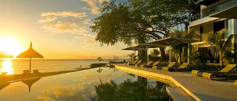 Un área tranquila junto a la piscina con sillones de descanso y sombrillas, con vista a una masa de agua en calma al atardecer.
