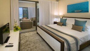 Habitación de hotel con una cama grande, televisión, escritorio con control remoto y vista al océano a través de una puerta abierta.