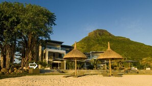 Un resort en la playa con montañas al fondo, que cuenta con un edificio moderno, sombrillas de paja y una playa de arena.