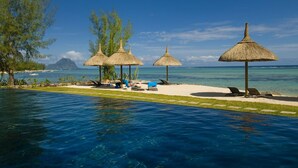 Zona junto a la piscina con sillones de descanso y sombrillas de paja, con vistas al mar azul y una montaña distante.