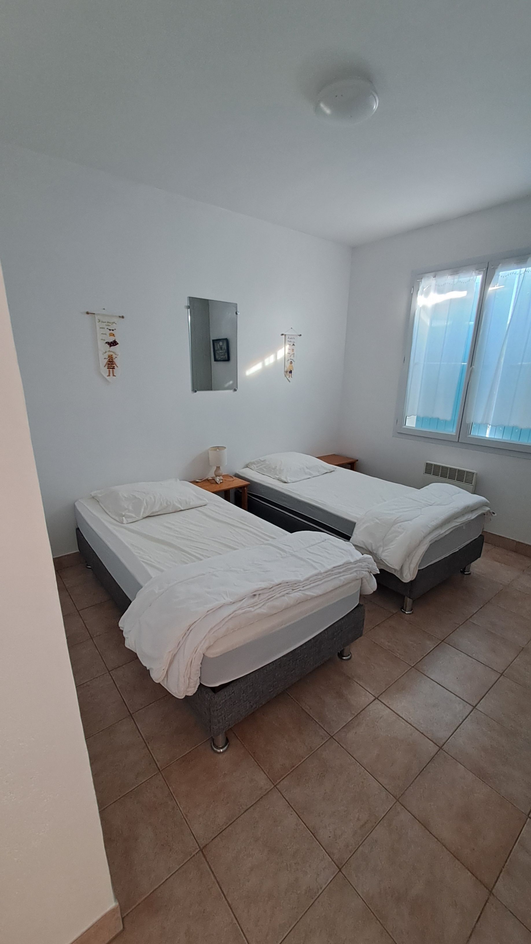 A room with two beds, a window, and a wall-mounted light fixture.