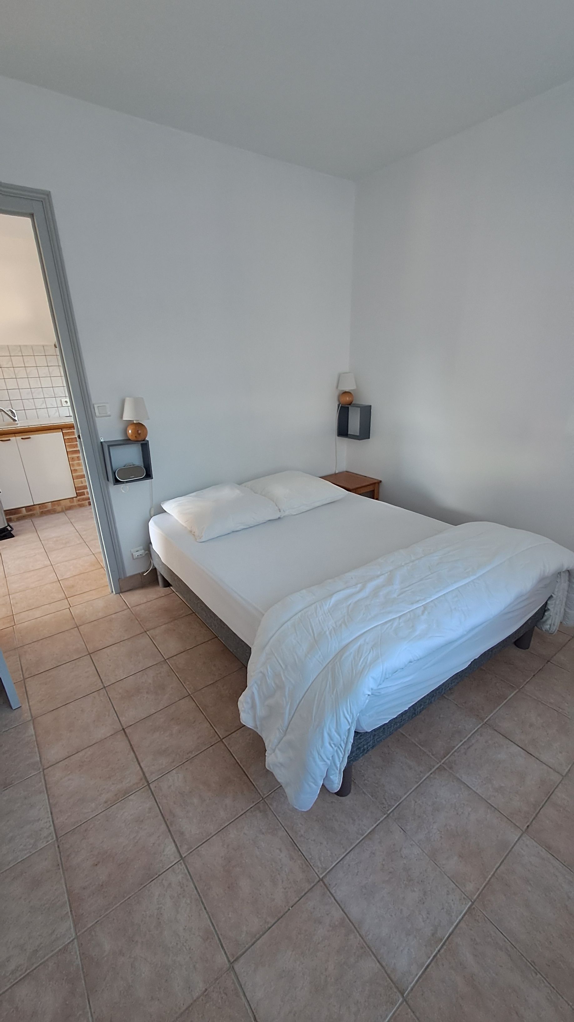 Une chambre à coucher avec un lit, une table de chevet, une lampe fixée au mur, et une salle de bain visible à travers une porte ouverte.