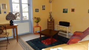 A living room with a sofa, a coffee table, a lamp, a television, and a window with curtains. - Colorful house in a green hamlet (Forgès)