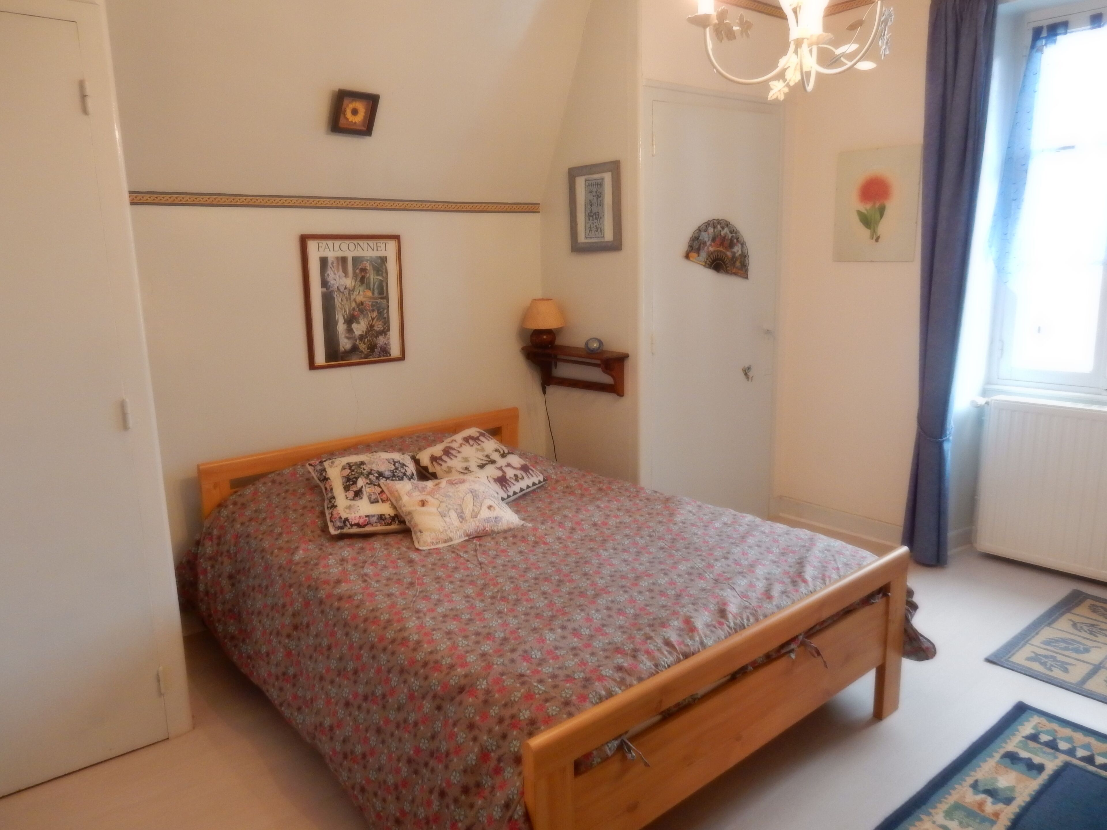 A bedroom with a wooden bed, floral bedding, a small bedside table with a lamp, and a picture frame on the wall.