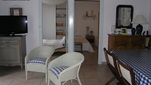 Une pièce comprenant un lit, deux fauteuils en osier, un téléviseur, une table recouverte d’une nappe à carreaux, un miroir et une étagère avec des objets décoratifs.