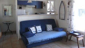 Un espace de vie compact comprenant un coin cuisine, un canapé avec des coussins, une petite table et une fenêtre avec des rideaux.