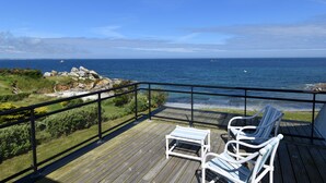 Terraza de madera con dos sillas blancas y una pequeña mesa, con vistas a la costa y el mar, con rocas a lo lejos.