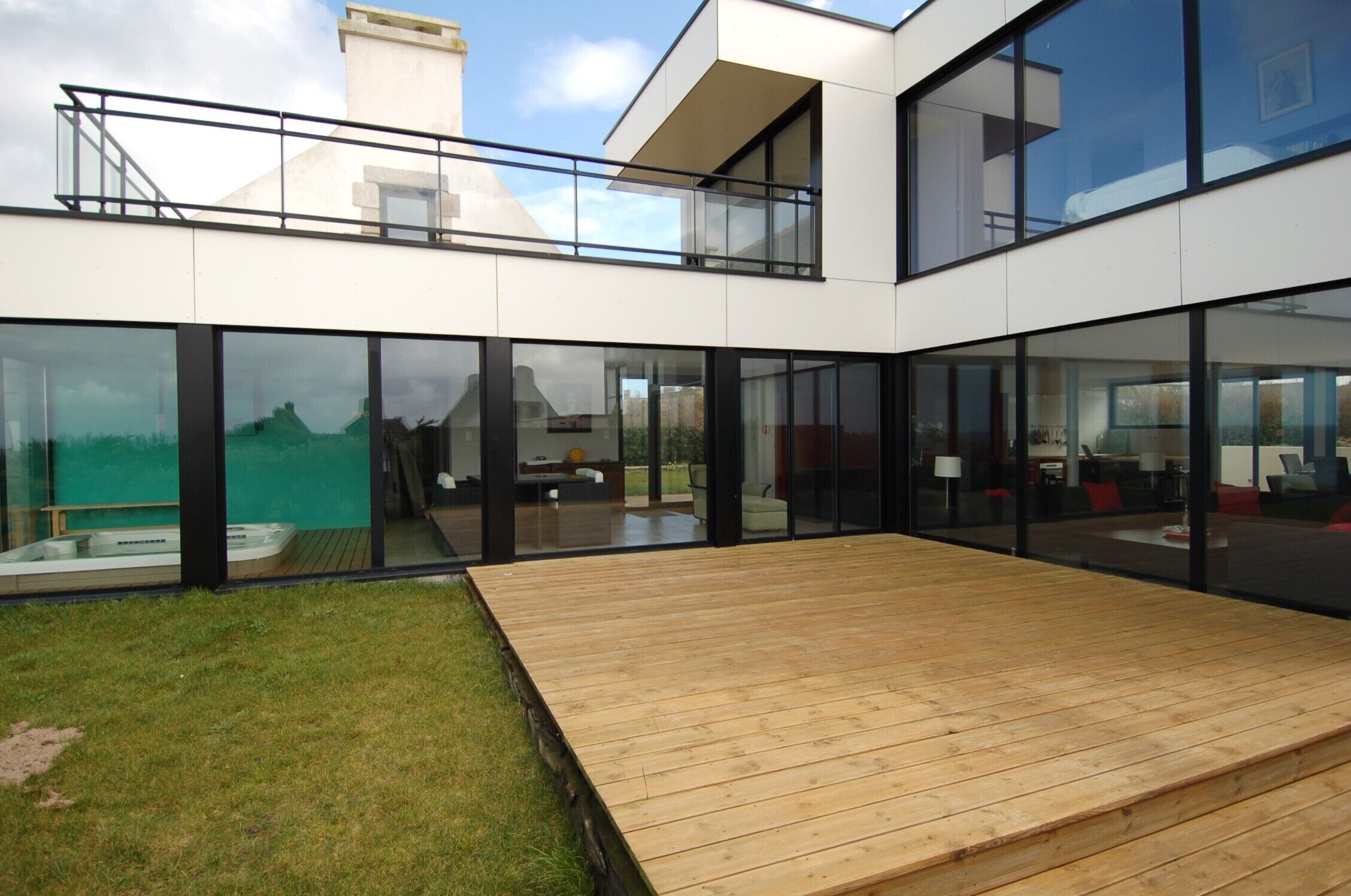 A modern house with large glass windows, a wooden deck, and a visible hot tub.