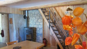 Ein rustikaler Innenraum mit Steinwand, einer Holztreppe, einem Esstisch und einem Kaminofen.