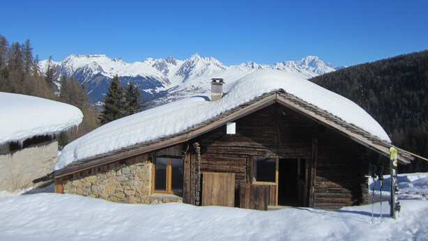 Eine Holzhütte mit Steinunterbau, umgeben von schneebedeckten Bergen und Bäumen.