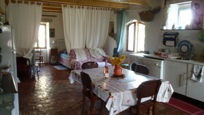 A rustic room with a bed, a table with chairs, a window with curtains, and a kitchen area.