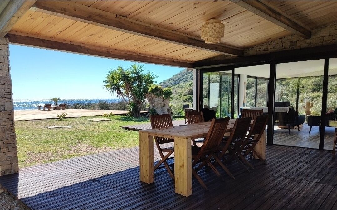 A wooden deck with a table and chairs, overlooking a scenic view of the sea and hills.
