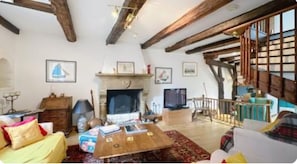 A cozy living room with a fireplace, wooden beams, a television, and a staircase leading to an upper level.