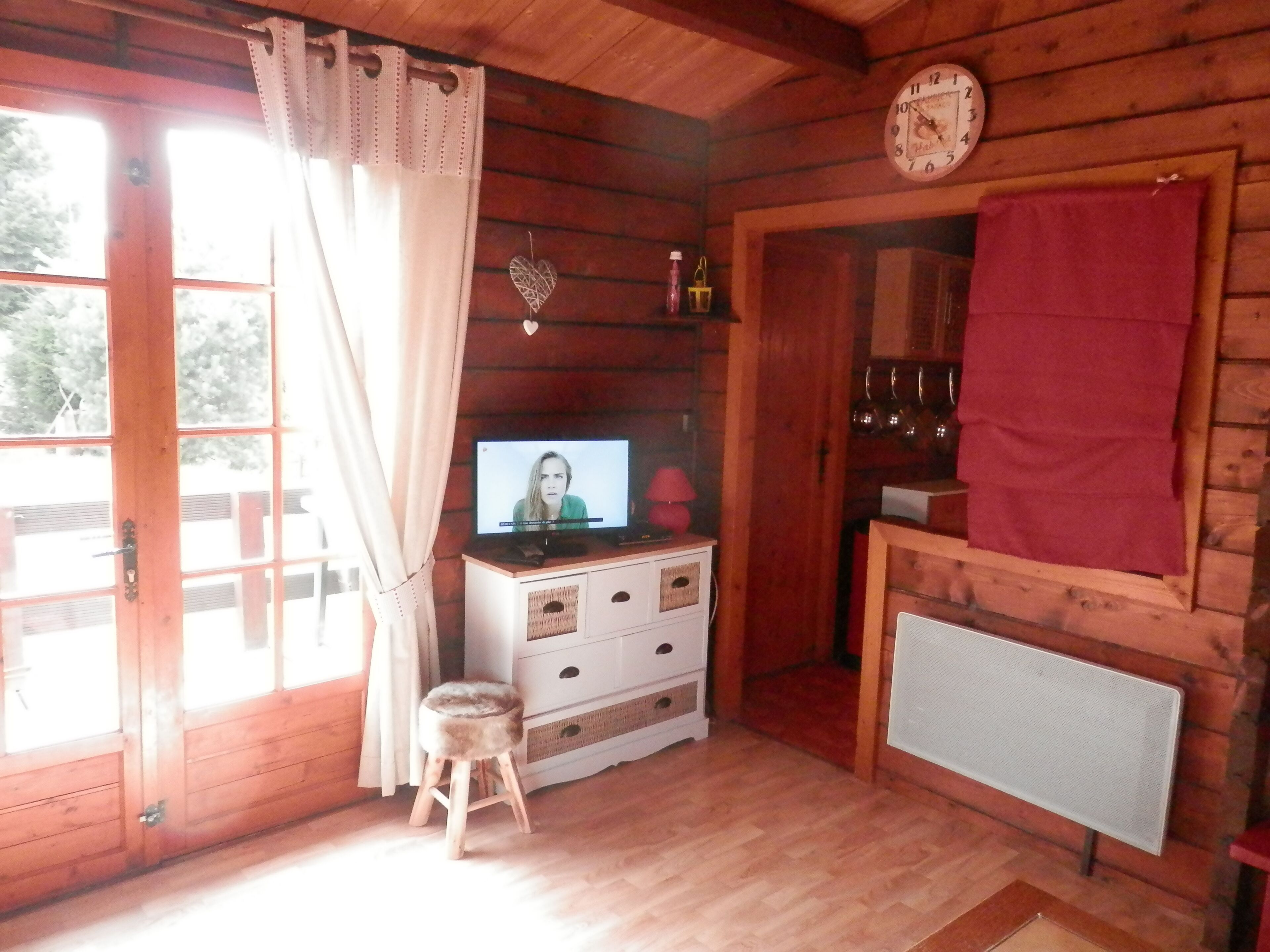 A cozy cabin room with a wooden interior, a television on a dresser, a clock on the wall, and a window with curtains.
