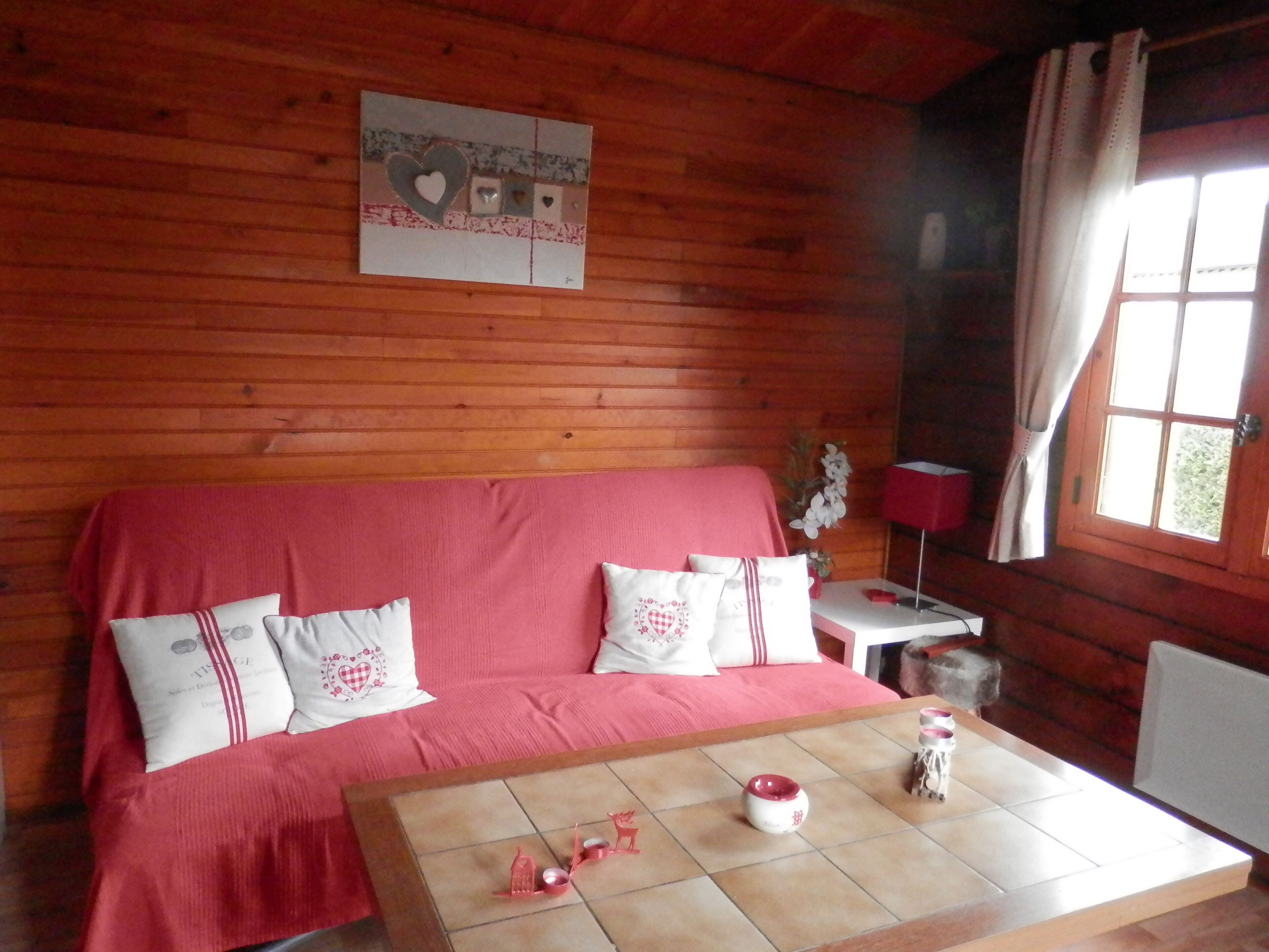 A cozy room with a wooden interior, a pink sofa with decorative pillows, a small table with a red lamp, and a window with curtains.