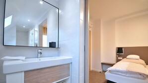 Ein modernes Hotelzimmer mit einem großen Spiegel, einem weißen Waschbecken, einer Holzkommode, einem Bett mit weißer Bettwäsche und einem Nachttisch mit Lampe.