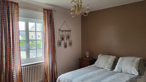 Une chambre à coucher avec un lit, une fenêtre avec des rideaux, un radiateur, une table de chevet avec une lampe et des tableaux accrochés au mur.